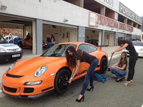 Flat bottomed girls in heels attempt to change tires on an orange Porsche 911 GT3